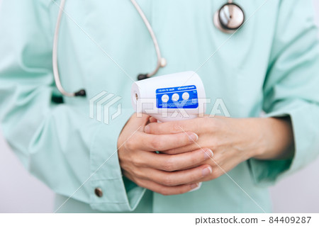 Temperature measurement gun in doctor hands. Close-up shot of doctor wearing protective surgical mask ready to use infrared isometric thermometer gun to check body temperature for virus symptoms. Temperature measurement gun in doctor hands. Close-up shot of doctor wearing protective surgical mask ready to use infrared isometric thermometer gun to check body temperature for virus symptoms. 84409287