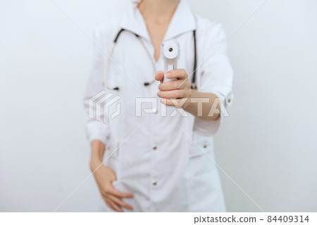 Temperature measurement gun in doctor hands. Close-up shot of doctor wearing protective surgical mask ready to use infrared isometric thermometer gun to check body temperature for virus symptoms. 84409314