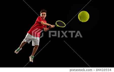Full-length portrait of boy, tennis player in motion, kicking ball with racket isolated over black backgound Full-length portrait of boy, tennis player in motion, kicking ball with racket isolated over black backgound 84410554