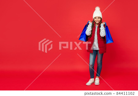 smiling kid in winter hat hold shopping bags on red background with copy space, closeout 84411102