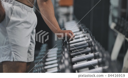 Arab man athlete is grabbing a dumbbell from a big selection in a fitness center. 84416919