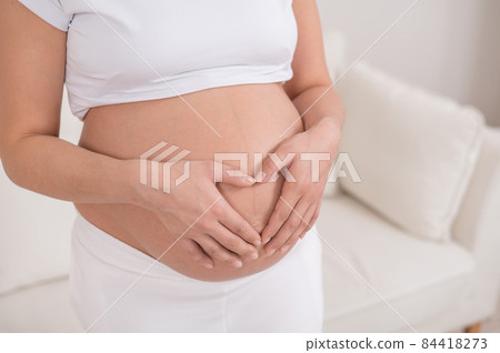 Close-up of a pregnant woman holding her hands in the shape of a heart on her stomach. 84418273