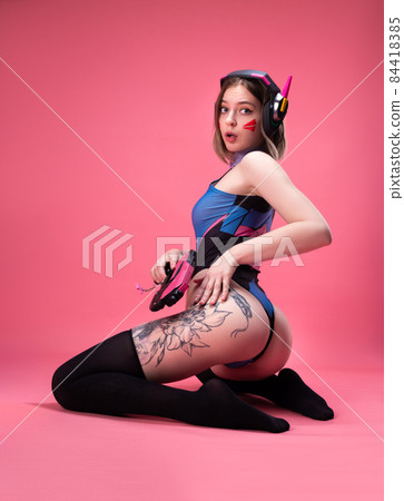 sexy gamer girl in a candid bright bodysuit and a gaming headset with a gun in her hands on a pink background 84418385