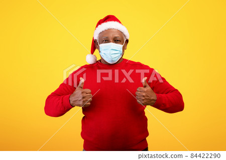 Senior black man in face mask and Santa hat showing thumbs up gesture with both hands on orange studio background 84422290