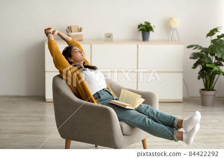 Carefree arab woman stretching in cozy armchair while reading and spending free time at home, having peaceful weekend Carefree arab woman stretching in cozy armchair while reading and spending free time at home, having peaceful weekend 84422292