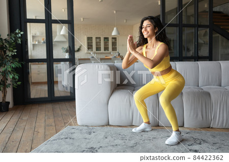 Full length of cheerful young fit woman doing squats, working out at home during covid lockdown, copy space 84422362