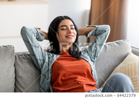 Relaxed young Arab woman leaning back on couch, resting with hands behind head on comfortable sofa, closing her eyes 84422379