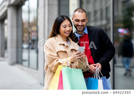Positive young diverse couple with bright paper bags and cellphone ordering goods online near shopping mall Positive young diverse couple with bright paper bags and cellphone ordering goods online near shopping mall 84422393