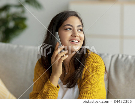 Cheerful middle eastern lady making call on mobile phone, sitting on sofa at home and looking aside at copy space Cheerful middle eastern lady making call on mobile phone, sitting on sofa at home and looking aside at copy space 84422396