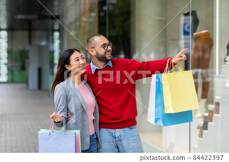 Affectionate young interracial couple standing on street with shopping bags, pointing at store window near city mall Affectionate young interracial couple standing on street with shopping bags, pointing at store window near city mall 84422397