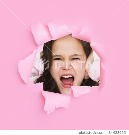 Frustrated sad european teen girl screams and covers her ears with hands looking through hole on pink paper Frustrated sad european teen girl screams and covers her ears with hands looking through hole on pink paper 84422633