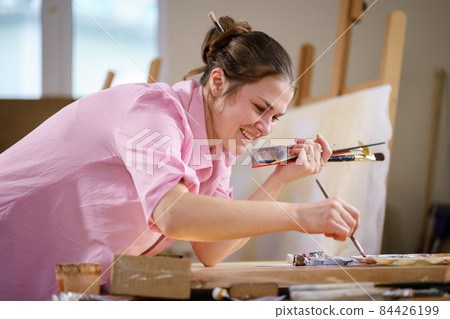 Cute woman paints on canvas in an art workshop. Artist creating picture. Art school or studio. Work with paints, brushes and easel. Hobby and leisure concept. Woman painter at workspace Cute woman paints on canvas in an art workshop. Artist creating picture. Art school or studio. Work with paints, brushes and easel. Hobby and leisure concept. Woman painter at workspace 84426199