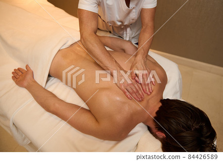 Overhead view of unrecognizable professional massage therapist massaging back muscles of a young man, relaxing, lying down on massage table at luxury wellness spa center. Alternative therapy concept 84426589