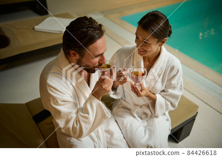 Beautiful loving couple of European man and woman, in white bathrobes, chilling, sitting close to each other by jacuzzi in luxurious wellness spa resort, quietly talking over cup of healthy tea drink Beautiful loving couple of European man and woman, in white bathrobes, chilling, sitting close to each other by jacuzzi in luxurious wellness spa resort, quietly talking over cup of healthy tea drink 84426618