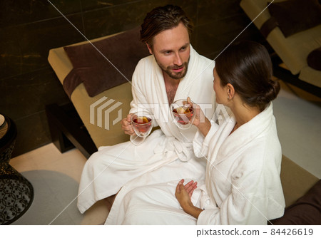 Handsome attractive man and beautiful gorgeous woman, both of them wearing white cozy terry bathrobes, resting in lounge zone by pool in luxury wellness spa centre, cutely talking over cup of tea Handsome attractive man and beautiful gorgeous woman, both of them wearing white cozy terry bathrobes, resting in lounge zone by pool in luxury wellness spa centre, cutely talking over cup of tea 84426619