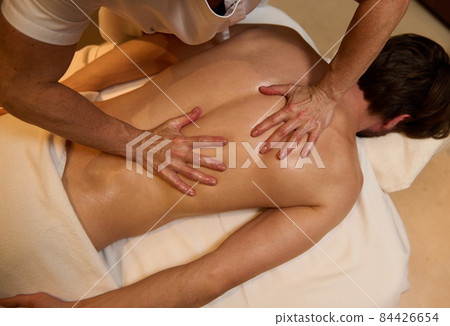 Overhead view of unrecognizable professional masseuse doing therapeutic healing massage of back muscles of a young man, relaxing on massage table at wellness spa centre. Alternative therapy concept 84426654