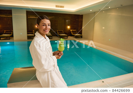 Charming woman in white bathrobe stands by the pool in luxurious a wellness spa complex, holds freshly squeezed vitamin juice in hands, smiles looking at camera. Healthy lifestyle, recreation concept Charming woman in white bathrobe stands by the pool in luxurious a wellness spa complex, holds freshly squeezed vitamin juice in hands, smiles looking at camera. Healthy lifestyle, recreation concept 84426699
