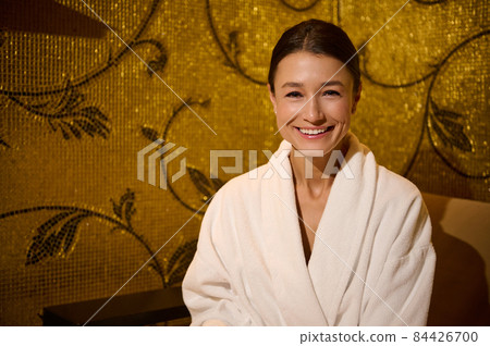 Close-up portrait of a beautiful stunning delighted middle aged European woman in white terry bathrobe smiling toothy smile looking at camera, sitting on an armchair in wellness spa centre lounge area Close-up portrait of a beautiful stunning delighted middle aged European woman in white terry bathrobe smiling toothy smile looking at camera, sitting on an armchair in wellness spa centre lounge area 84426700