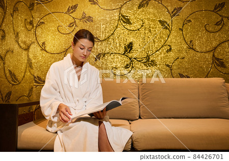 Confident well-groomed middle aged woman in white terry bathrobe reading magazine sitting in a couch in a lounge area of a luxury wellness spa centre. Professional body, skin care, relaxation concept Confident well-groomed middle aged woman in white terry bathrobe reading magazine sitting in a couch in a lounge area of a luxury wellness spa centre. Professional body, skin care, relaxation concept 84426701