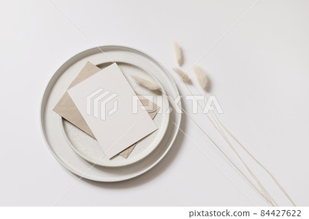 Modern autumn stationery still life. Blank greeting card mock up scene with Lagurus ovatus grass, craft envelope and ceramic plates isolated on white table background. Flat lay, top view, no people. Modern autumn stationery still life. Blank greeting card mock up scene with Lagurus ovatus grass, craft envelope and ceramic plates isolated on white table background. Flat lay, top view, no people. 84427622