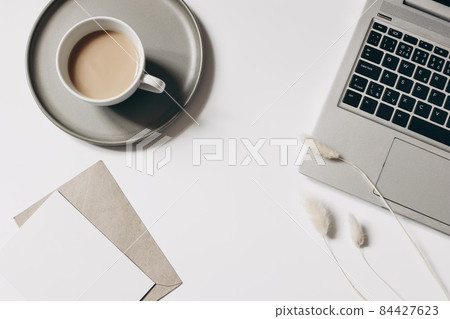 Elegant workspace composition. Cup of coffee, dry lagurus grass and laptop isolated on white table background. Blank greeting card mockup with envelope. Flat lay, top view. Blogger, home office. Elegant workspace composition. Cup of coffee, dry lagurus grass and laptop isolated on white table background. Blank greeting card mockup with envelope. Flat lay, top view. Blogger, home office. 84427623