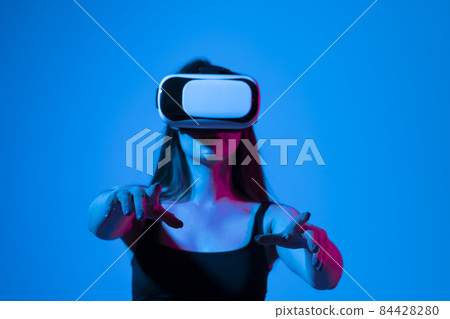 Cheerful girl with hands up wearing the virtual reality goggles and playing a games in neon light. Future technology concept. Metaverse. 84428280