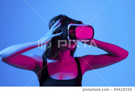 Cheerful girl with hands up wearing the virtual reality goggles and playing a games in neon light. Future technology concept. Metaverse. 84428359