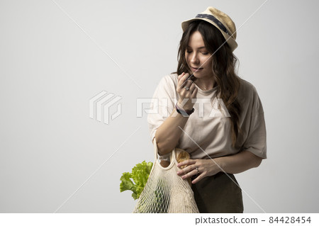 Happy woman in a beige t-shirt and a hat holding reusable cotton shopping bag with groceries, bread and greens. Concept of no plastic. Zero waste, plastic free. Sustainable lifestyle. 84428454