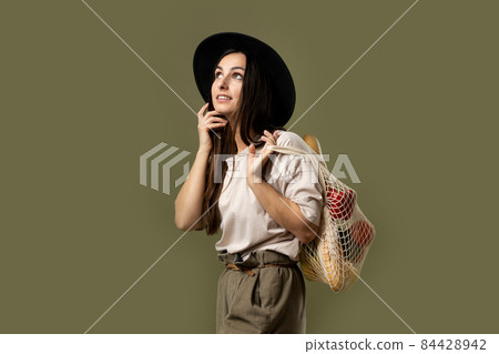 Woman in a beige t-shirt and a hat holding mesh shopping bag with groceries. Conscious consumption. Eco trend. Zero waste concept. Girl carry bag shopper. Woman in a beige t-shirt and a hat holding mesh shopping bag with groceries. Conscious consumption. Eco trend. Zero waste concept. Girl carry bag shopper. 84428942