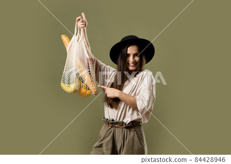 Mesh eco bag with groceries, vegetables and fruits, in the hands of a brunette girl in a beige t-shirt and black hat. Eco friendly concept. Woman holding string shopping bag. Mesh eco bag with groceries, vegetables and fruits, in the hands of a brunette girl in a beige t-shirt and black hat. Eco friendly concept. Woman holding string shopping bag. 84428946