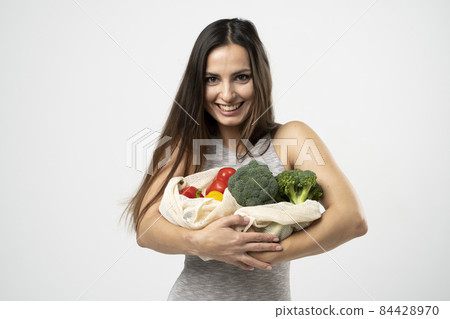 A white mesh mesh bag with groceries in a hands of a girl in grey dress against the white background of a studio. 84428970