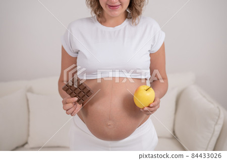 A pregnant woman is holding an apple and a bar of chocolate. The ninth month of pregnancy. A pregnant woman is holding an apple and a bar of chocolate. The ninth month of pregnancy. 84433026