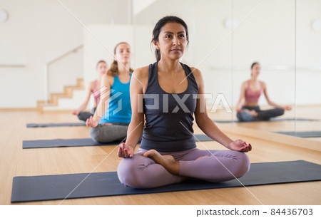 Concentrated Latina sitting in lotus pose with group of women 84436703