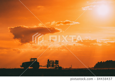 Tractor Rides On Countryside Road. Beginning Of Agricultural Spring Season. Cultivator Pulled By A Tractor In Rural Field Landscape Under Sunny Summer Sunset Sunrise Sky. Backlit Dramatic Lighting Tractor Rides On Countryside Road. Beginning Of Agricultural Spring Season. Cultivator Pulled By A Tractor In Rural Field Landscape Under Sunny Summer Sunset Sunrise Sky. Backlit Dramatic Lighting 84438437