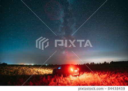 Night Starry Sky With Glowing Stars Above Countryside Road Landscape With SUV Car Vehicle Automobile. Milky Way Galaxy And Rural Field Meadow 84438439