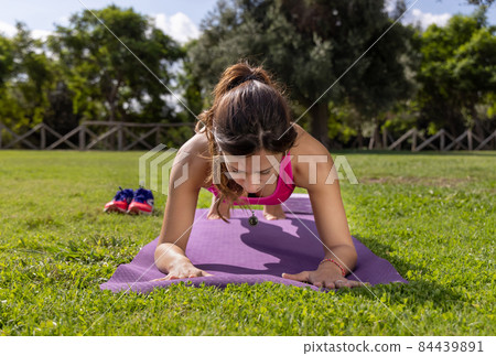 Young concentrated woman in plank position on mat in a park in a summer day Young concentrated woman in plank position on mat in a park in a summer day 84439891