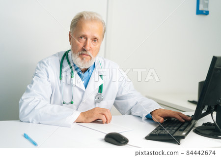 Portrait of serious mature adult male doctor in white coat with stethoscope working on desktop computer at desk in medical office, looking at camera. 84442674