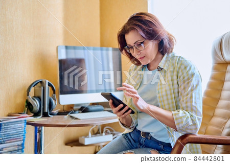 Middle aged woman using smartphone sitting in armchair in home office Middle aged woman using smartphone sitting in armchair in home office 84442801