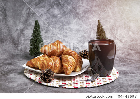 Cup of hot chocolate with a plate with croissant on a Christmas tablecloth and some Christmas fir trees and pine cones Cup of hot chocolate with a plate with croissant on a Christmas tablecloth and some Christmas fir trees and pine cones 84443698
