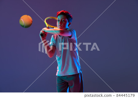 Motivated, concentrated boy training, playing tennis, kicking ball with racket isolated over purple background in neon lights 84445179