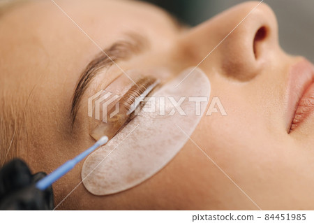 Close-up of eyelash care treatment procedures. Beautiful female, blond hair model have eyelashes lamination. Woman doing staining, curling, laminating and extension for lashes 84451985