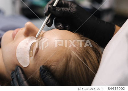 Close-up of eyelash care treatment procedures. Beautiful female, blond hair model have eyelashes lamination. Woman doing staining, curling, laminating and extension for lashes Close-up of eyelash care treatment procedures. Beautiful female, blond hair model have eyelashes lamination. Woman doing staining, curling, laminating and extension for lashes 84452002