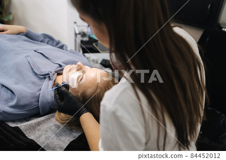 Woman doing eyelashes lamination in beauty studio. Blond hair model has staining, curling, laminating and extension for lashes. Black gloves Woman doing eyelashes lamination in beauty studio. Blond hair model has staining, curling, laminating and extension for lashes. Black gloves 84452012