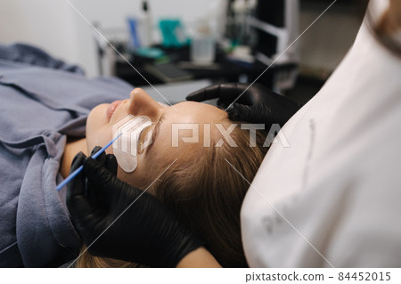 Woman doing eyelashes lamination in beauty studio. Blond hair model has staining, curling, laminating and extension for lashes. Black gloves Woman doing eyelashes lamination in beauty studio. Blond hair model has staining, curling, laminating and extension for lashes. Black gloves 84452015