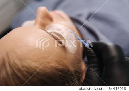 Close-up of eyelash care treatment procedures. Beautiful female, blond hair model have eyelashes lamination. Woman doing staining, curling, laminating and extension for lashes Close-up of eyelash care treatment procedures. Beautiful female, blond hair model have eyelashes lamination. Woman doing staining, curling, laminating and extension for lashes 84452018