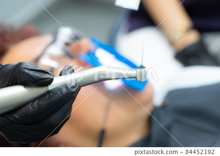 Drill with a tool in the hands of a dentist in black gloves for the treatment of pulpitis, canals in dentistry. Against the background of a patient with glasses Drill with a tool in the hands of a dentist in black gloves for the treatment of pulpitis, canals in dentistry. Against the background of a patient with glasses 84452192