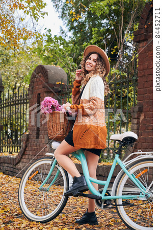 Side view of curly brunette travelling by vintage cycle with big basket of pink flowers. Pretty female wearing brown hat and cardigan is riding bike in autumn park. Concept of retro style. 84452711
