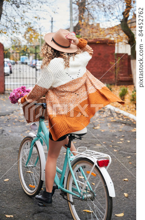 Rear view of biker riding on sidewalk by bright women's bicycle, holding hat on head with her right hand. On the background brick wall with gate, blurred silhouettes of cars. 84452762