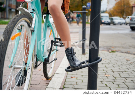 Close up view of female legs on light blue bike in retro style near bicycle parking rack at city street. Active recreation while riding on cycle. Close up view of female legs on light blue bike in retro style near bicycle parking rack at city street. Active recreation while riding on cycle. 84452784