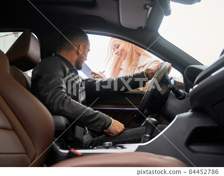 Young handsome happy man customer sitting inside new car. Beautiful smiling woman salesperson asking to sign some important documents at car dealership outdoors. View from inside a car. 84452786